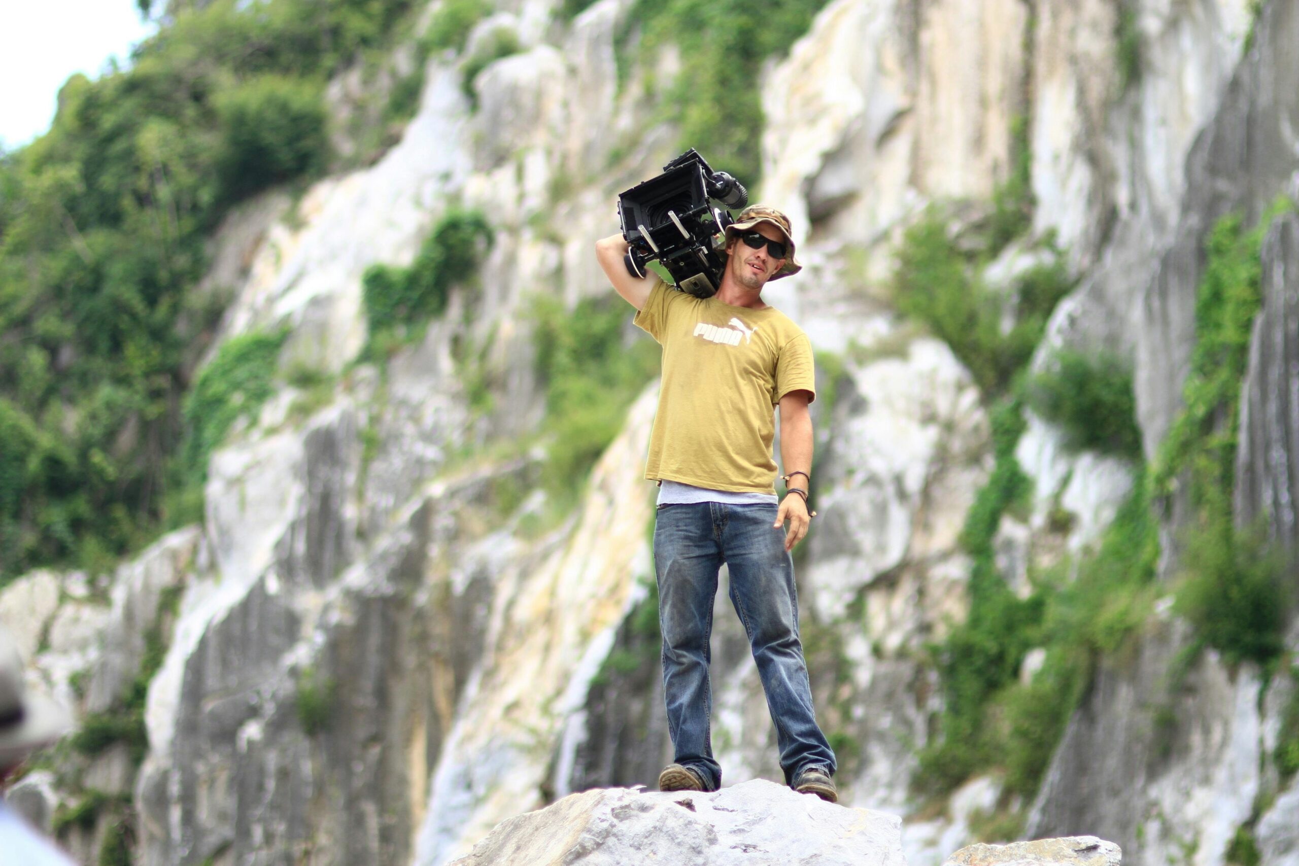 A cinematographer holding a camera stands on a rocky cliff, capturing stunning outdoor footage.